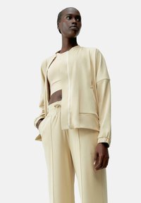 Beige sportos szett, amely tartalmaz egy cipzáros dzsekit, crop toppot és magas derekú nadrágot. Simaság jellemzi, finom varrási részletekkel és zsebekkel.