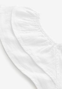 Vit kortärmad topp med volangkanter, med en rynkad textur och en subtil sömdetalj längs halslinningen. Lättviktsmaterial.