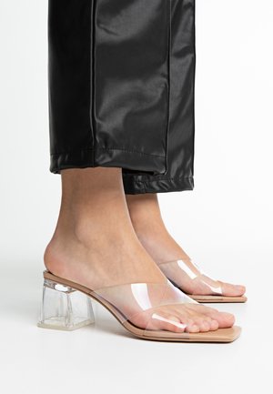 Transparent block-heeled sandals with nude footbed, featuring a clear plastic strap design and open toe, set against black pants.