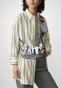 Μαύρη και λευκή τσάντα με καμπύλο σχήμα, που διαθέτει έντονη γραφή και αλυσίδα. Φορεμένη με ριγέ oversized πουκάμισο και γκρίζα παντελόνια.