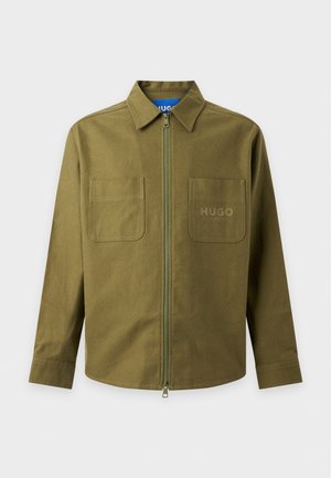Jeune homme debout, mains dans les poches, portant une veste zippée vert olive, un pantalon cargo noir et des baskets foncées sur fond blanc.