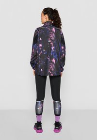 Veste de sport noire avec un imprimé abstrait coloré, associée à des leggings noirs avec des accents assortis et des chaussettes texturées violettes.