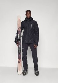 Άνδρας με μαύρο μπουφάν σκι, παντελόνι και μπότες κρατώντας ένα όρθιο σκι σε απλή ανοιχτή φόντο.