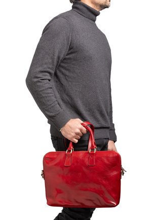 Homme en col roulé gris tenant une mallette en cuir rouge brillant par ses poignées sur fond blanc.