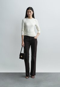 Λευκό ριμπ πλεκτό μακρυμάνικο τοπ, σκούρο καφέ παντελόνι, μικρή σκοτεινή τσάντα, πέδιλα ανοιχτής μύτης· μινιμαλιστικός σχεδιασμός με εφαρμοστή σιλουέτα.