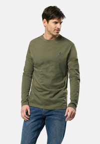 Chemise à manches longues vert olive en coton, avec un col rond et un petit logo bleu sur la poitrine, assortie à un jean bleu.