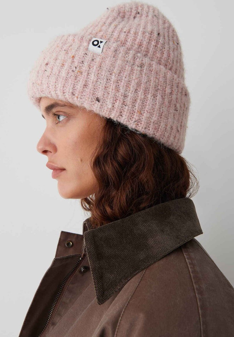 Roze gebreide beanie met een geribbelde textuur en een klein label, gedragen met een bruine jas met een corduroy kraag.