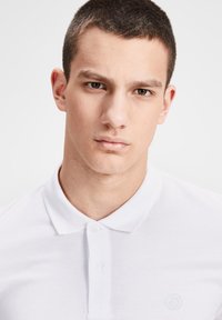 Polo shirt blanc en tissu texturé, doté d'un col classique et d'une fermeture à deux boutons. Petit logo sur le côté gauche de la poitrine.