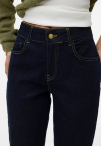Donkerblauwe denim jeans met goudkleurige stiksels, een voorste knoopsluiting en zijzakken, die een getailleerd ontwerp en een gladde textuur laten zien.