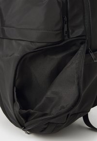 Dettaglio di uno zaino nero con tasca con zip, realizzato in tessuto liscio con una finitura opaca, che mostra una fodera interna e dettagli di cucitura.