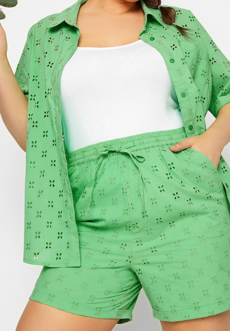 Femme portant un short vert à œillets et une chemise assortie ouverte sur un débardeur blanc, la main dans la poche, tenant des lunettes de soleil.