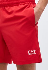 Άνδρας που φοράει κόκκινα σορτς EA7 Emporio Armani με ελαστική μέση, χέρι στην τσέπη, κοντινό πλάνο στο λογότυπο στο δεξί μηρό.