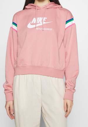 Femme portant un sweat à capuche de sport Nike rose avec des rayures multicolores sur les manches et un pantalon beige clair, debout devant un fond blanc.