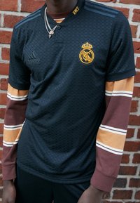 Černý strukturovaný dres s tlačným límcem, zdobený zlatým logem Real Madrid, a dlouhými rukávy s hnědými, zlatými a bílými pruhy.