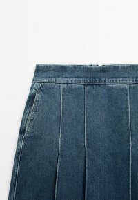 Jupe en jean bleu avec des détails de coutures verticales et une poche latérale, ceinture visible, posée sur un fond blanc uni.
