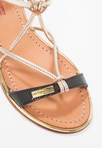 Sandalo con cinturini caratterizzato da accenti metallici dorati, soletta in pelle marrone e un cinturino testurizzato nero con una piastra con logo.