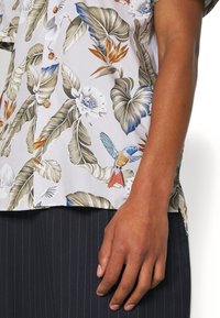 Camicetta a maniche corte con uno sfondo grigio chiaro, caratterizzata da motivi colorati di foglie e fiori tropicali, e un accento di uccello blu.