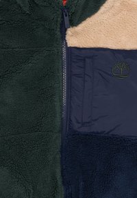 Fleece-jakke med hette, med mørkegrønne og marineblå seksjoner. Tan fleece-detalj på den øvre delen og glidelås foran. Brodert logo.
