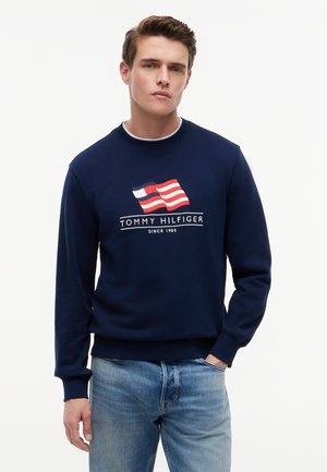 Άνδρας που φοράει ναυτικό μπλε φούτερ Tommy Hilfiger με λογότυπο σημαίας σε κόκκινο, άσπρο και μπλε, και ανοιχτό μπλε τζιν, στέκεται με το ένα χέρι στην τσέπη.