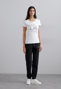 T-shirt bianco a maniche corte con logo EA7 nero, abbinato a pantaloni jogger neri. Modello indossa sneakers bianche, in piedi su uno sfondo neutro.