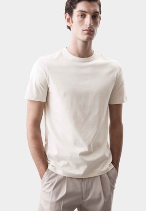 Hombre joven con una camiseta blanca de manga corta lisa y pantalones beige claros, de pie con las manos en los bolsillos frente a un fondo neutro.