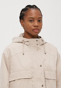 Beige huppari, jossa on nappi- ja vetoketjukiinnitys, kahdella etutaskulla, säädettävillä nyöreillä ja sileällä, matalla pinnalla.