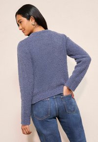 Moteris vilki purų violetinį megztinį ir mėlynas džinsus, pozuoja viena ranka kišenėje, atsukusi nugarą į kamerą.