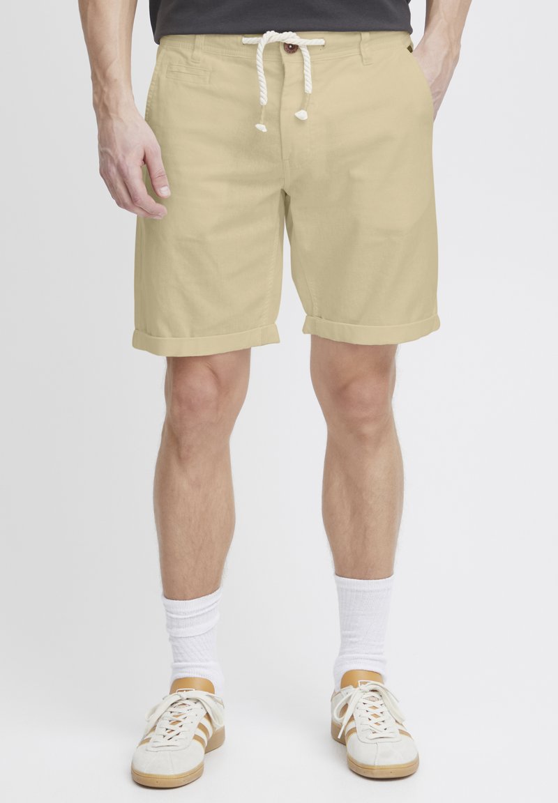 Shorts en coton beige avec des revers roulés, taille avec cordon de serrage et détail de bouton. Associés à des chaussettes blanches et des baskets légères.