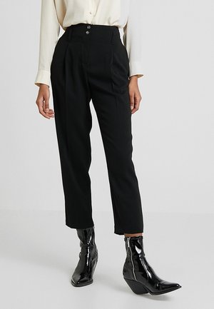 Femme portant un pantalon noir taille haute avec plis devant et détail de bouton, assorti de bottines noires brillantes et d’un chemisier clair.