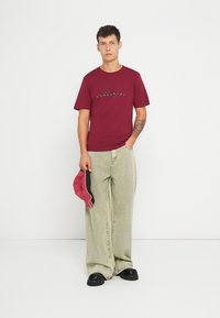 Tricou bordeaux cu text grafic negru, asortat cu pantaloni lejeri de culoare verde deschis. Modelul ține o șapcă roșie și neagră și poartă pantofi negri.