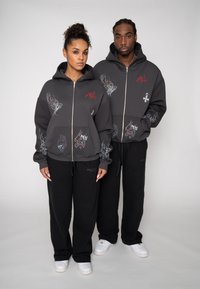 Dunkelgraue Kapuzenjacken mit Reißverschluss und grafischen Designs, kombiniert mit schwarzen Jogginghosen. Das Material wirkt weich, mit Fronttaschen und gerippten Bündchen.