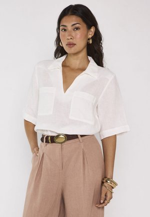 Femme debout face à l'avant portant un chemisier blanc à manches courtes, un pantalon plissé beige taille haute, une ceinture marron, des bijoux dorés et des boucles d’oreilles créoles.