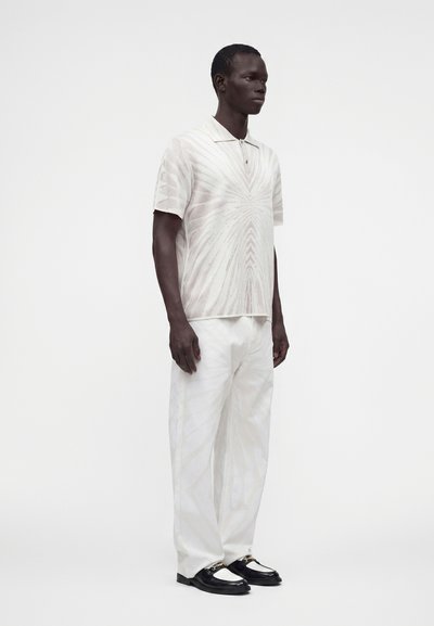 Homme debout portant un polo à manches courtes à motif clair, un pantalon blanc ample et des mocassins noirs et blancs sur un fond uni.
