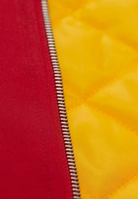 Roter und gelber Stoffreißverschluss-Detail, das eine gesteppten Textur zeigt, mit einem glänzenden Metallreißverschluss, der entlang der Naht verläuft.