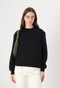 Felpa nera con collo a girocollo a costine e polsini elasticizzati. Abbinata a jeans bianchi con cuciture in vista. Tracolla verde su borsa a spalla.