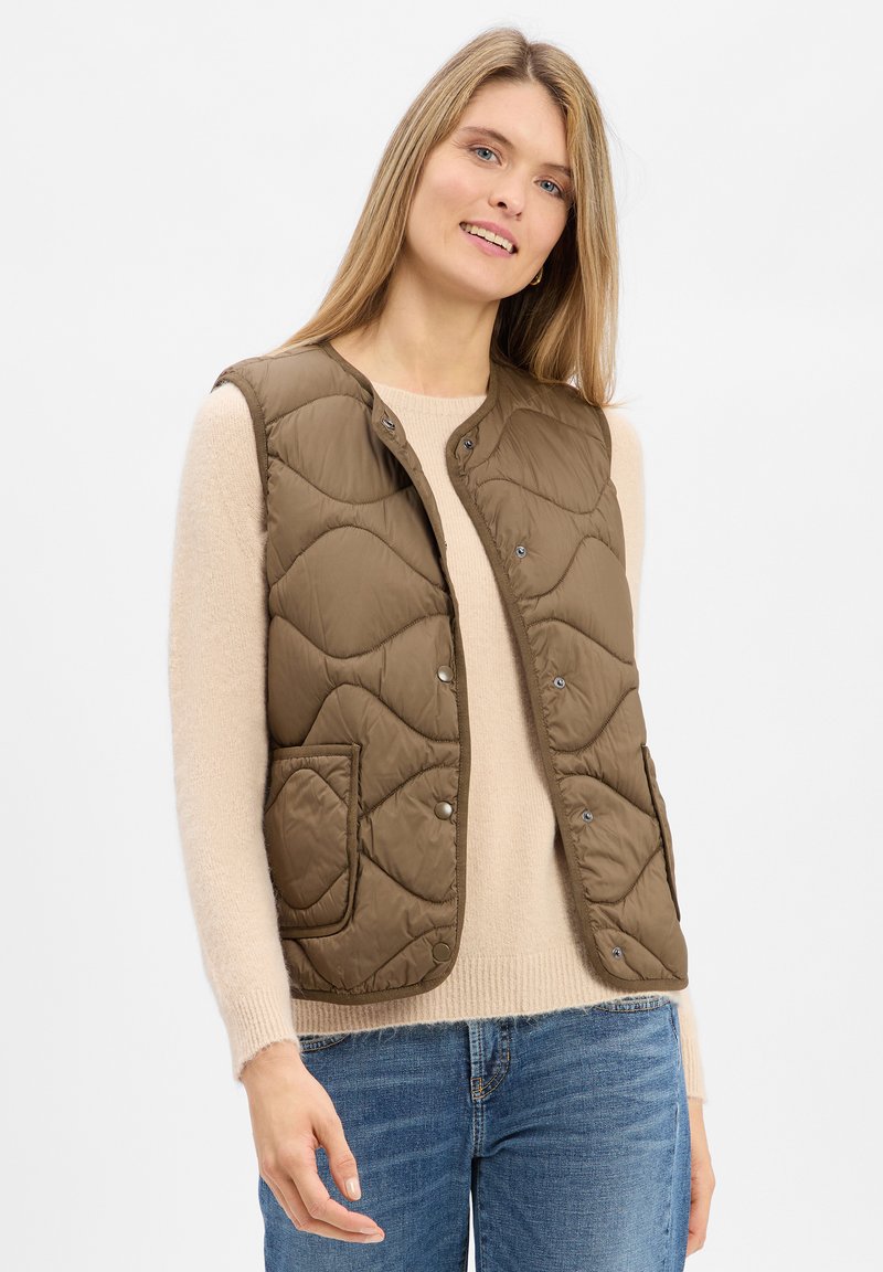 Frau in einer braunen gesteppte ärmellosen Weste über einem beigen Pullover und blauen Jeans, stehend vor einem schlichten weißen Hintergrund.