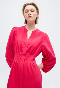 Νεαρή γυναίκα με κοντά ξανθά μαλλιά που φοράει ένα φωτεινό ροζ φόρεμα με φουσκωτά μακριά μανίκια, πόζα σε απλό φωτεινό φόντο.