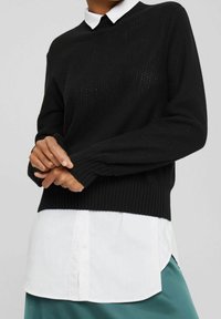 Zwarte textuur lange mouwen trui over een witte colbertshirt, gedragen door een persoon met de handen voor zich gekruist, met een turquoise rok zichtbaar onderaan.