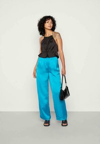 Mulher com cabelo encaracolado, vestindo uma blusa preta sem mangas e calças largas azul brilhante, segurando uma bolsa preta, de pé contra um fundo liso.