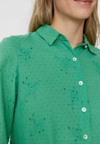 Camicia verde con bottoni, realizzata in tessuto testurizzato, con ricami ad occhiello e piccoli bottoni circolari, dotata di colletto e disegni floreali.