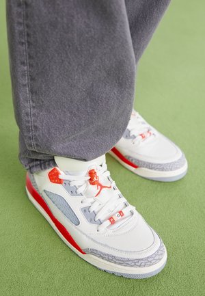 Osoba nosząca białe, czerwone i szare sneakersy z oznaczeniem PSG oraz szare spodnie, stojąca na zielonym dywanie.