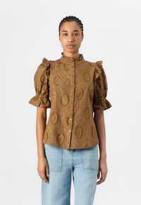 Blusa marrone con maniche a palloncino, dettagli arricciati e un motivo ornamentale. Presenta un colletto alto e una chiusura con bottoni, abbinata a jeans azzurri.