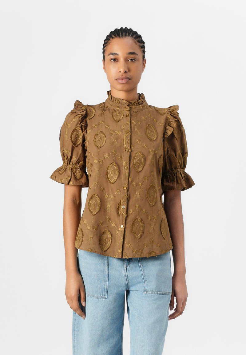 Blusa marrone con maniche a palloncino, dettagli arricciati e un motivo ornamentale. Presenta un colletto alto e una chiusura con bottoni, abbinata a jeans azzurri.