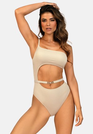 Kobieta o długich falowanych włosach, ubrana w beżowy kostium kąpielowy na jedno ramię z dużym wycięciem i paskiem w talii, pozująca z uniesioną jedną ręką.