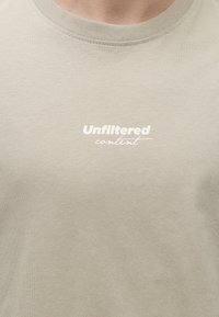 Κοντινή λήψη ενός μπεζ T-shirt με λευκό κείμενο που λέει "Unfiltered content" στο στήθος.