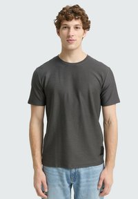 Tricou scurt de culoare gri închis, fabricat dintr-un material moale, cu o guler rotund și o textură subtilă, purtat cu blugi de un albastru deschis.