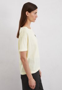 Geel T-shirt met korte mouwen en een ronde halslijn, voorzien van een klein zwart logo op de borst, gemaakt van zachte, lichte stof.