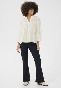 Blusa a righe semi-trasparente di colore chiaro con colletto; maniche lunghe a sbuffo. Abbinata a jeans a zampa d'elefante blu scuro e scarpe color crema.