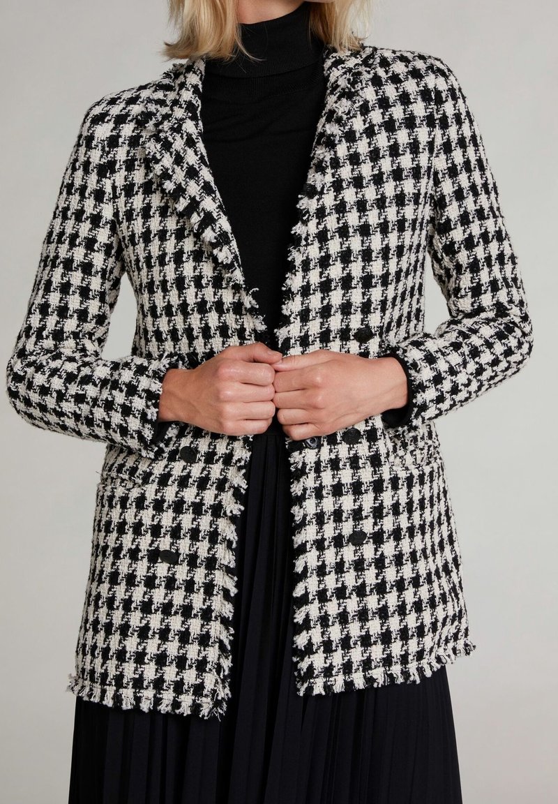 Blazer à motif pied-de-poule noir et blanc avec un bord frangé, deux poches avant, fermeture à bouton et un col, porté par-dessus un pull col roulé noir.