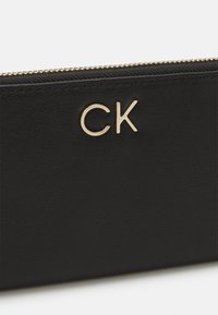 Melna ādas maks ar teksturētu virsmu, kas aprīkots ar zelta "CK" logo un zelta rāvējslēdzēja aizdari.